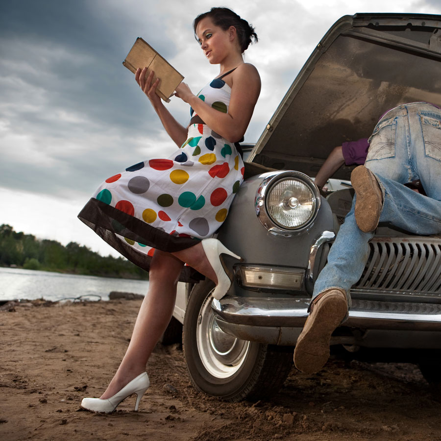 Eine Frau liest ein Buch während ein Mann versucht, das Auto zu reparieren